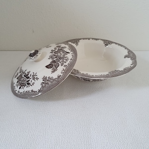 Pode incluir: Uma tigela de servir de cerâmica branca com um padrão floral castanho e uma tampa correspondente. A tigela tem uma borda ondulada e a tampa tem um botão no topo.