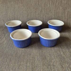 Può includere: Un set di cinque piccole pirofile in ceramica rotonde, caratterizzate da un esterno blu vivace con dettagli a coste verticali e un interno liscio color bianco sporco. Ogni contenitore presenta un bordo largo e piatto e una forma circolare.