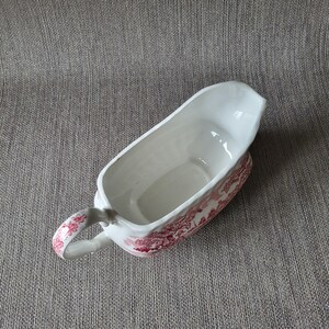 May include: A white ceramic gravy boat with a red floral pattern. The boat has a handle and a spout for easy pouring. The gravy boat is approximately 8 inches long.