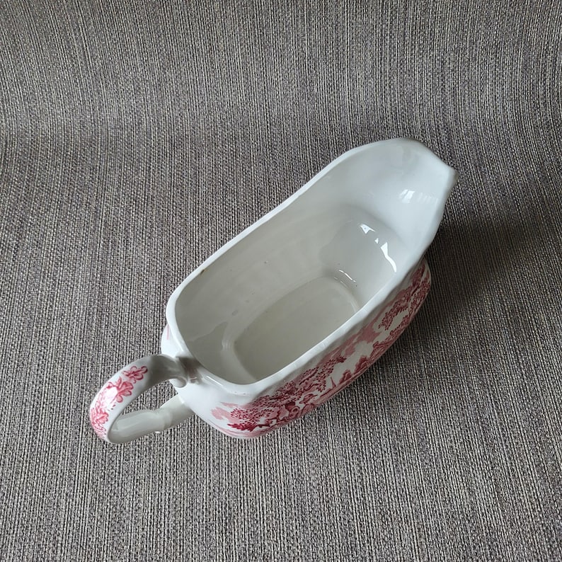 May include: A white ceramic gravy boat with a red floral pattern. The boat has a handle and a spout for easy pouring. The gravy boat is approximately 8 inches long.