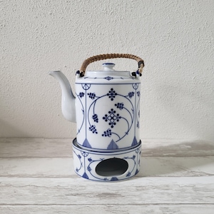 Puede incluir: Una tetera blanca con un diseño floral azul y blanco y un asa tejida. La tetera está sentada sobre una base blanca y azul a juego con un agujero en el centro.