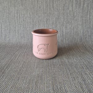 May include: A small, cylindrical, light pink ceramic cup with a smooth finish. The exterior features an embossed line drawing of a stylised animal and the text SAN SALVATORE. The interior of the cup is a contrasting dark terracotta brown colour.