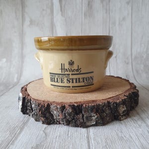 May include: A small, cream-colored ceramic crock with brown trim and two handles. The crock has a black label that reads "Harrods Finest Selected Blue Stilton Cheese".