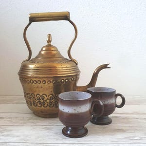 Könnte beinhalten: Eine Messing-Teekanne mit einem verzierten Design und zwei braunen und weißen Keramikbechern.