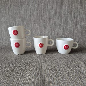 Puede incluir: Cuatro tazas de espresso de cerámica blanca con una pequeña asa en el lateral. Cada taza presenta un emblema de sello de cera circular rojo en la parte frontal con las letras blancas D-E impresas en el centro. Dos tazas están apiladas.