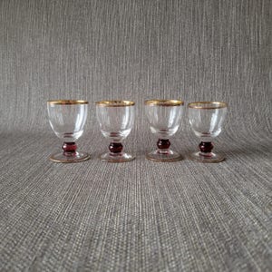 Può includere: Quattro bicchieri da liquore in vetro trasparente con bordi dorati e steli rosso rubino. I bicchieri sono disposti in fila su una superficie grigia strutturata. I bicchieri hanno un design classico ed elegante.
