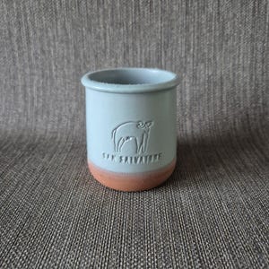 Può includere: Un bicchiere in ceramica azzurra con base color terracotta. Il bicchiere presenta un'immagine in rilievo di una mucca e la scritta "SAN SALVATORE". La tazza cilindrica ha una consistenza leggermente ruvida.