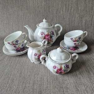 Peut inclure: Un service à thé en porcelaine blanche comprenant une théière avec couvercle, un pot à lait, un sucrier avec couvercle et deux tasses avec soucoupes assorties. Chaque pièce est ornée d'un motif floral coloré de fleurs roses, violettes et jaunes.