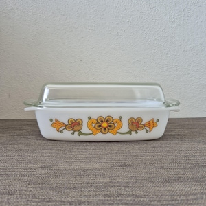 Puede incluir: Una fuente rectangular de vidrio blanco con tapa de vidrio transparente. El exterior de la fuente presenta un patrón floral repetitivo en tonos naranja, amarillo y verde oliva, con flores y hojas estilizadas y simétricas en el centro.