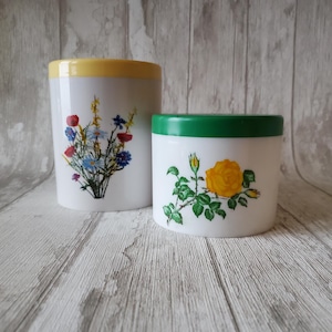 Puede incluir: Dos recipientes cilíndricos blancos con tapas. Uno tiene una tapa amarilla y un diseño de ramo floral. El otro tiene una tapa verde y un diseño de rosa amarilla. Los recipientes están sobre una superficie de madera clara.