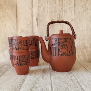 Puede incluir: Una tetera de terracota con asa trenzada y un juego de seis tazas a juego. La tetera y las tazas están decoradas con un intrincado diseño de animales y personas.