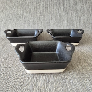 Op de afbeelding: Drie vierkante ovenschaaltjes met elk twee handgrepen aan de zijkant. De schaaltjes hebben een matzwarte binnenkant en bovenkant, met een contrasterende witte onderkant. Het ontwerp is eenvoudig, functioneel en heeft een stevige vorm.