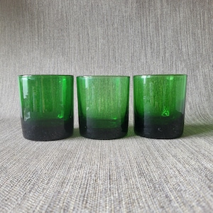 May include: Three green drinking glasses with a dark green base. The glasses are cylindrical and appear to be made of glass. The background is a textured grey fabric.
