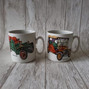 Puede incluir: Dos tazas de cerámica blanca con imágenes de coches antiguos. La taza de la izquierda tiene un Cadillac verde de 1908. La taza de la derecha tiene un Ford Modelo T rojo de 1908.