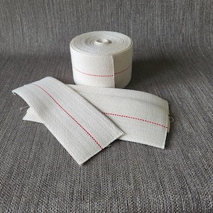 May include: A roll of white cotton wick with a red stripe, and two pieces of the same wick laid out. The wick is tightly woven and appears to be new. The background is a textured gray fabric.
