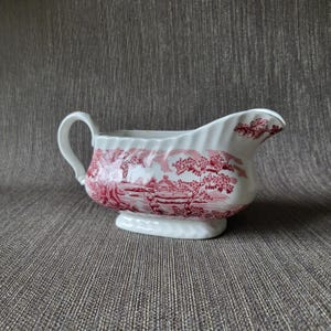 May include: White ceramic gravy boat with a red transferware pattern depicting a landscape scene. The boat has a handle and a spout, and sits on a small, oval base. The design is a classic, vintage style.