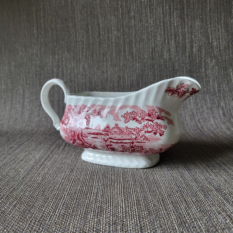 May include: White ceramic gravy boat with a red transferware pattern depicting a landscape scene. The boat has a handle and a spout, and sits on a small, oval base. The design is a classic, vintage style.