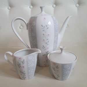 Puede incluir: Un juego de té de porcelana blanca con tetera, jarra de crema y azucarero. El juego tiene un patrón de rayas grises con detalles florales de rosas rosas y hojas verdes. La tetera tiene un diseño alto y delgado.