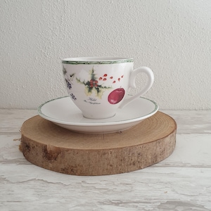 Può includere: Un set di tazza e piattino bianchi con bordo verde e un design decorativo con una mela, agrifoglio e bacche. Il set è posto su una fetta di legno, creando un'estetica rustica.