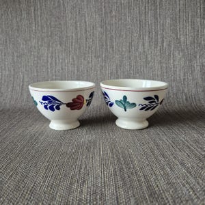 May include: Two white ceramic bowls with a floral design. The bowls have a footed base and a red rim. The floral design includes blue, red, and green leaves and stems. The bowls are set against a gray background.
