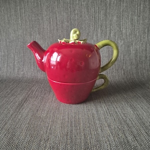 Può includere: Servizio da tè in ceramica rossa a forma di fragola. La teiera ha un manico, coperchio e foglie verdi. La tazza rossa è impilata sotto, creando un design unico.