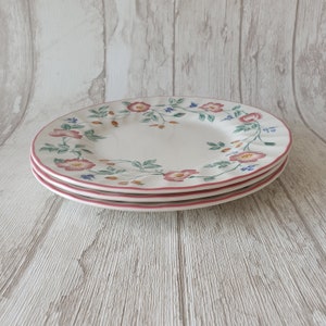 May include: Three white ceramic plates with a pink floral design. The plates have a scalloped edge and are stacked on top of each other.