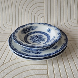 May include: A stack of three white ceramic plates featuring a blue floral pattern. The design includes a central rose motif, a decorative lattice border, and a scalloped edge with blue detailing. The plates are layered, showing the intricate blue and white transferware style on each piece.