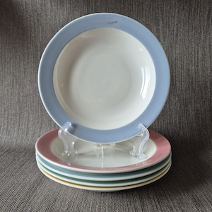 Puede incluir: Una pila de platos de cerámica con un centro blanco y un borde de color. El plato superior tiene un borde azul claro, y los platos de abajo tienen bordes rosa, turquesa y amarillo. La palabra "California" está impresa en el plato superior.