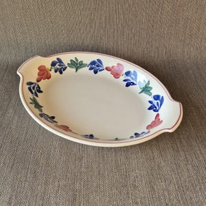 May include: Oval serving platter with a white base and a floral design. The rim is outlined in red, and the flowers are in shades of blue, pink, and green. The platter has two handles and is approximately 14 inches long.