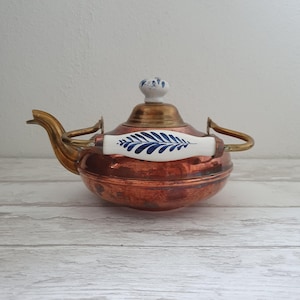 Może przedstawiać: Karafka do herbaty wykonana z miedzi z mosiężnym dzióbkiem i uchwytem. Karafka ma białą porcelanową pokrywkę z niebieskim wzorem kwiatowym i biały porcelanowy uchwyt z niebieskim wzorem kwiatowym.