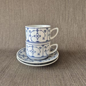 Puede incluir: Dos tazas de té de porcelana blanca apiladas, cada una con un asa y un diseño floral azul. Dos platillos a juego debajo. Las tazas y platillos tienen una estética clásica y elegante.