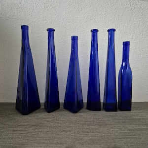Könnte beinhalten: Eine Sammlung von sechs kobaltblauen Glasflaschen in unterschiedlichen Höhen und Formen. Fünf der Flaschen haben einen markanten dreieckigen Boden, der sich zu einem schmalen Hals verjüngt, während die sechste Flasche einen zylindrischen Boden hat.