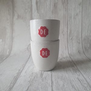 May include: Two white speckled ceramic plant pots stacked. Each pot features a red floral design with the letters "D-E". The pots are on a light grey and white wooden surface. The pots are suitable for indoor plants.