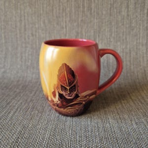 May include: A ceramic coffee mug with a red handle and interior. The exterior features a warrior illustration in shades of red, yellow, and brown. The mug has a glossy finish and is set against a neutral background.
