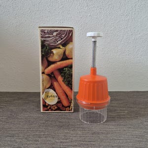 May include: A Zyliss brand manual food chopper with an orange plastic top, a white plunger handle, a metal spring, and a clear plastic base. The chopper is positioned next to its original cardboard box, which features images of vegetables.