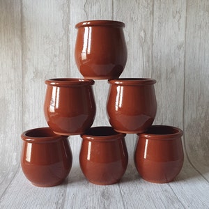 May include: Six small, brown ceramic pots stacked in a pyramid shape. The pots have a rounded shape with a slightly flared rim. The pots are a solid, glossy brown color.