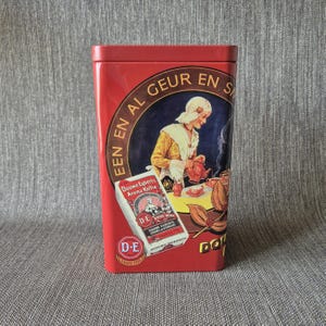 Op de afbeelding: Een rechthoekig rood metalen blik met een vintage illustratie van een persoon die koffie inschenkt. De tekst luidt EEN EN AL GEUR EN SMAAK en Douwe Egberts Aroma Koffie. Een klein D-E logo staat linksonder. Het blik heeft een deksel.