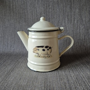 Puede incluir: Tetera de esmalte color crema con borde y asa negros. La tetera presenta una ilustración pintada de un cerdo con manchas negras. La tapa tiene un pequeño pomo. El pico está en el lado izquierdo.