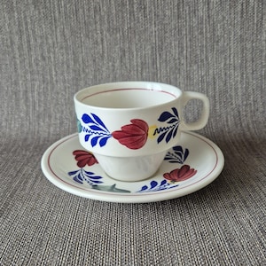 Könnte beinhalten: Weißes Tee-Set mit Blumendesign. Die Tasse und Untertasse haben rote, blaue und gelbe Blumenmuster. Der Rand der Tasse und Untertasse ist mit einer dünnen roten Linie verziert. Das Set steht auf grauem Hintergrund.