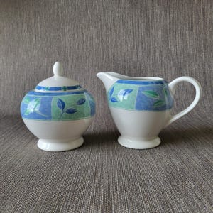Op de afbeelding: Een witte keramische suikerpot met deksel en een melkkan. Beide stukken hebben een blauw en groen bladeren ontwerp op een witte achtergrond. De suikerpot heeft een ronde vorm en een klein handvat op het deksel.