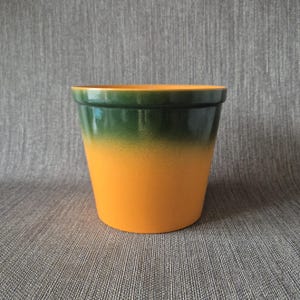 Puede incluir: Una maceta cilíndrica de cerámica con un acabado degradado suave que transiciona desde un verde bosque profundo en el borde superior hasta un naranja vibrante y sólido en la base. La maceta tiene una abertura ancha y una base plana.