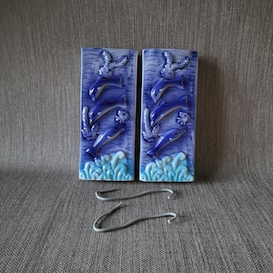 Könnte beinhalten: Zwei blaue Keramik-Wandplatten mit einem Reliefdesign von Delfinen, die durch Wellen springen. Jede Platte zeigt drei Delfine, Seetang und einen Seestern. Zwei silberne Metallhaken befinden sich darunter.