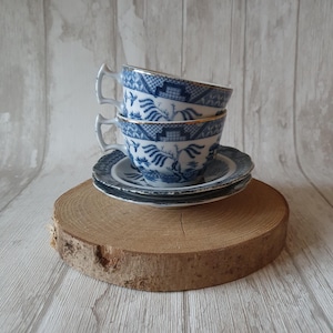 Puede incluir: Tres tazas de té azules y blancas con un borde dorado apiladas una encima de la otra sobre una base de madera. Las tazas tienen un diseño chino tradicional con un sauce llorón y pájaros.