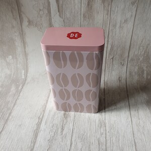 May include: A pink metal storage container with a white lid and a brown coffee bean pattern. The lid has a red circle with the letters "D-E" in white.