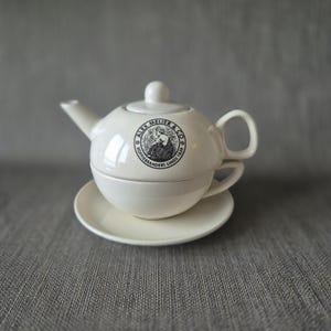 以下が含まれることがあります： 白のセラミックティーセット。ティーポット、ティーカップ、ソーサーが含まれています。ティーポットには注ぎ口と取っ手があり、ティーカップはティーポットの中に収まります。「ALEX MEIJER & CO」と書かれた白黒のロゴがあります。