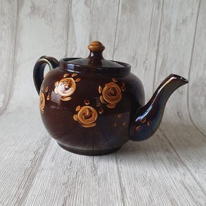Può includere: Una teiera in ceramica marrone con un motivo floreale. La teiera ha un coperchio e un beccuccio. Il motivo floreale è di colore marrone chiaro e presenta rose stilizzate.