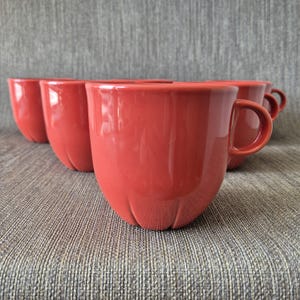 May include: A set of four red ceramic mugs. Each mug has a rounded body with a small handle and a unique, textured base. The mugs are arranged in a slightly offset pattern, showcasing their design.