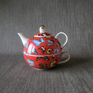 Puede incluir: Juego de tetera y taza rojo con un diseño de mariposas. La tetera tiene un pico y asa blancos, y un pomo dorado en la tapa. La taza está apilada debajo de la tetera, creando un diseño único.