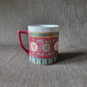 May include: A ceramic mug with a red handle and a white interior. The mug features a red background with floral and Chinese character designs. The top and bottom edges have colourful geometric patterns.