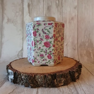 Puede incluir: Un tarro hexagonal de cerámica blanca con un patrón floral con rosas rosas y hojas verdes. El tarro está sentado sobre una rodaja de madera.